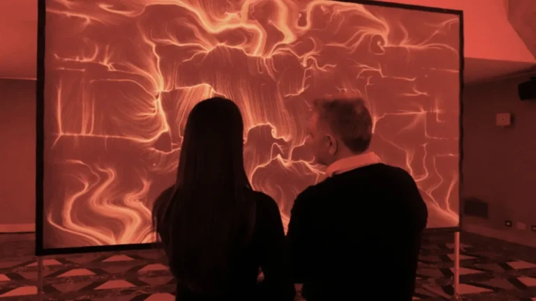 In una sala illuminata di rosso intenso, due persone di spalle, una con capelli lunghi scuri e una con capelli chiari, osservano un grande schermo montato su piantana. Sullo schermo un'opera dalle fitte linee e dai vortici rossi e dorati su fondo scuro. Il pavimento ha una decorazione geometrica a rombi; le pareti riflettono la luce rossa.