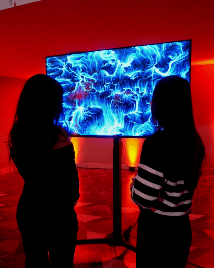 Due donne di spalle davanti a uno schermo su piantana che mostra un'opera dalle linee e dai vortici azzurri e blu; la sala è illuminata di rosso, con pavimento a scacchi e faretti a terra.