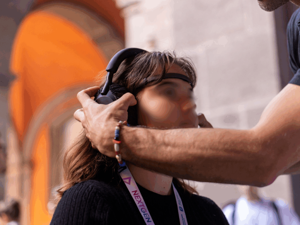 Un operatore sistema sulla testa di una partecipante seduta un archetto EEG sulla fronte e le cuffie; la ragazza indossa un pass NextGen e sullo sfondo si vedono le arcate di un edificio storico.