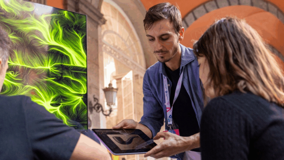 Un membro dello staff BrainArt® mostra un tablet a due partecipanti accanto a un'opera d'arte generativa dalle linee verdi su fondo scuro, sotto le arcate di un edificio storico.
