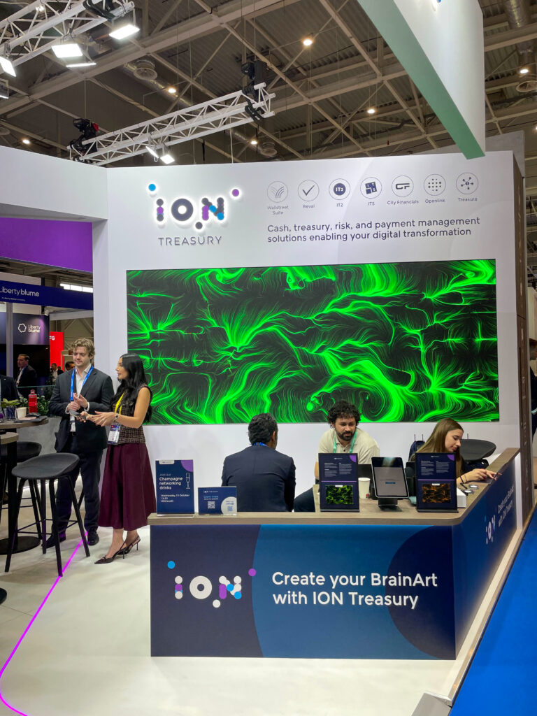 Stand fieristico con l'insegna "ION TREASURY" e il claim "Cash, treasury, risk, and payment management solutions enabling your digital transformation". Al centro, uno schermo a parete mostra un'opera dalle linee e dai vortici verdi su fondo scuro. Il bancone reception riporta la scritta "Create your BrainArt with ION Treasury"; dietro, alcune persone dello staff sedute e in piedi. In alto una struttura a traliccio con fari.
