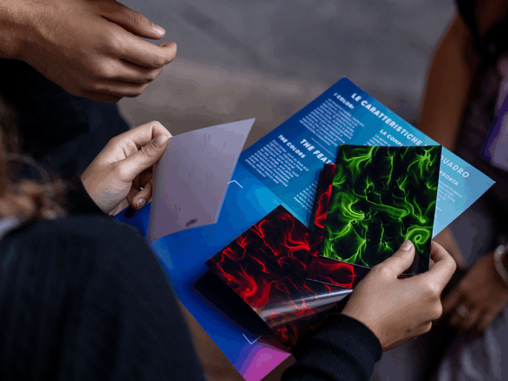 Mani che sfogliano un pieghevole BrainArt® con testo bilingue su fondo blu e alcune stampe di opere dalle linee e dai vortici, una rossa e una verde, su fondo nero.
