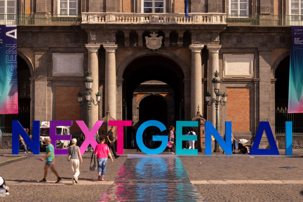 Grande installazione con le lettere colorate NEXTGEN AI davanti alla facciata monumentale in pietra del Palazzo Reale di Napoli, con persone che passeggiano sulla piazza.