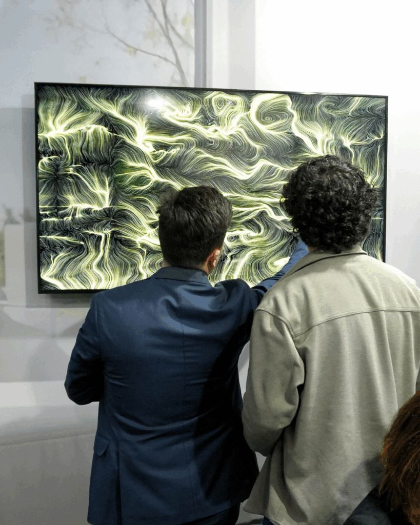 Due uomini di spalle, uno con giacca blu e uno con giacca chiara, osservano da vicino uno schermo a parete; quello a sinistra indica un punto dell'immagine. Sullo schermo un'opera dalle fitte linee e dai vortici in tonalità verdi e dorate chiare su fondo scuro. La parete intorno è chiara con una decorazione di rami sottili.