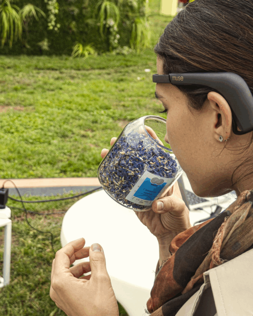 Una donna ripresa di profilo da dietro, all'aperto, con un sensore EEG scuro indossato sulla fronte e sopra le orecchie, avvicina al naso un barattolo di vetro pieno di fiori e petali essiccati azzurri e violacei, con un'etichetta sul fronte. L'altra mano è sollevata con le dita raccolte. Sullo sfondo un prato verde, una parete vegetale, un tavolino bianco con un cavo e un computer portatile.
