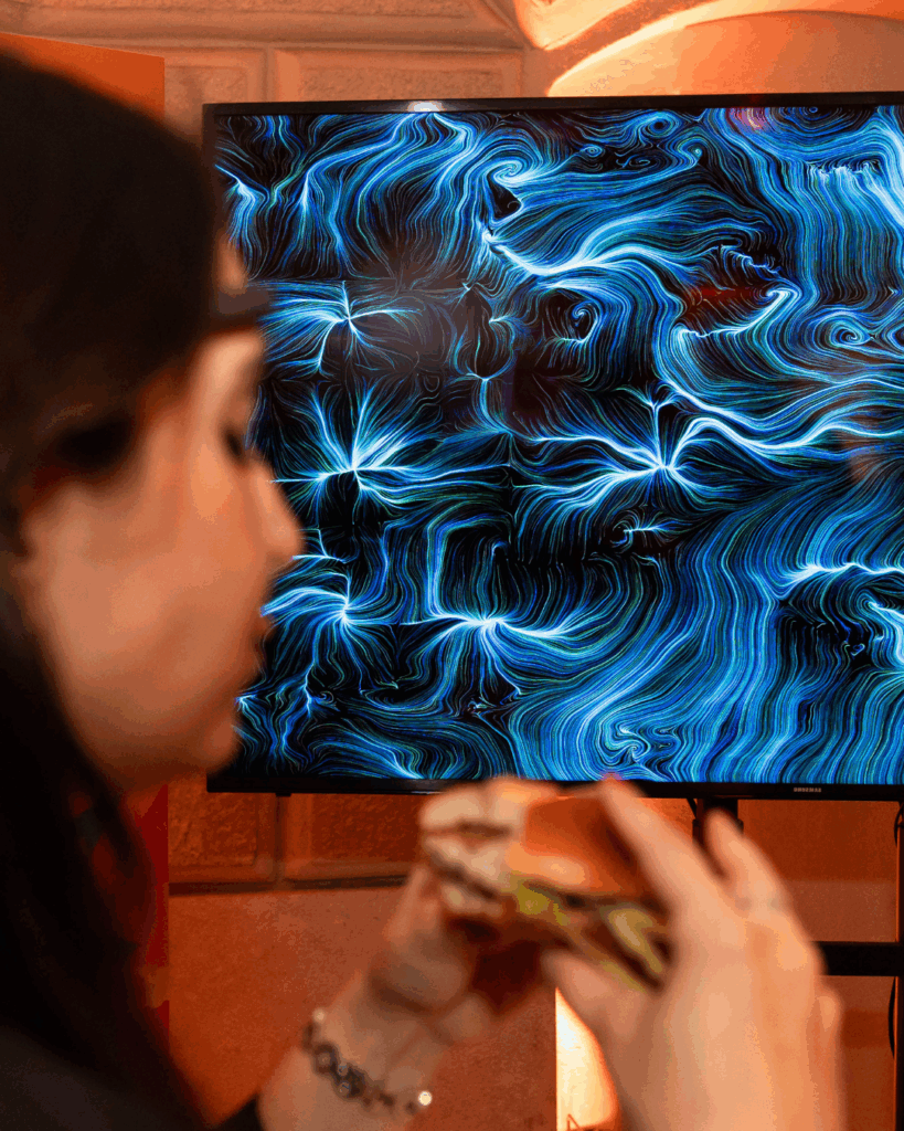In primo piano, di profilo e leggermente sfocata, una donna con capelli scuri tiene tra le mani un panino e osserva uno schermo. Sullo schermo un'opera dalle fitte linee e dai vortici blu e azzurri luminosi su fondo scuro, più brillanti in alcuni punti a raggiera. Sullo sfondo una parete chiara con luce calda.