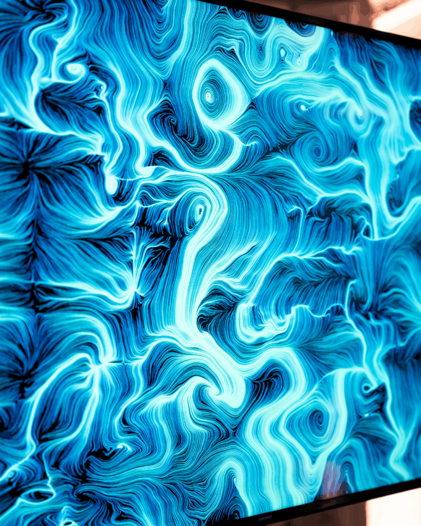 Primo piano ravvicinato di uno schermo ripreso in leggera diagonale, che mostra un'opera dalle fittissime linee azzurre e bianche luminose che si curvano e si intrecciano formando vortici su fondo blu scuro. La trama è densa e occupa tutta la superficie; sui bordi si intravede la cornice scura del display.