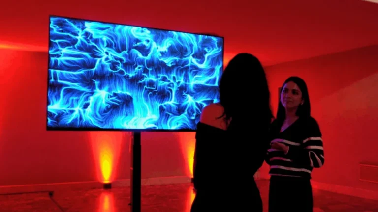 In una sala illuminata di rosso, due donne in primo piano osservano uno schermo su piantana: una è ripresa di spalle in silhouette, l'altra, girata di tre quarti, indossa un cardigan scuro a righe chiare. Sullo schermo un'opera dalle fitte linee e dai vortici blu su fondo scuro. Ai lati della base due fari a terra proiettano una luce arancione, il pavimento lucido ne riflette i bagliori e a terra corre un cavo.