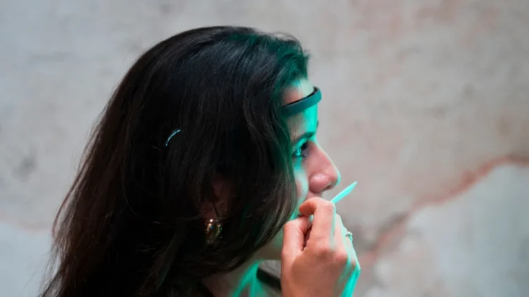 Una donna ripresa di profilo, capelli scuri sciolti, indossa sulla fronte una fascia scura con sensore e avvicina alle labbra una striscia da profumo (mouillette) che tiene con la mano destra. Indossa un top nero a spalline sottili, un filo rosso al polso e un anello. Il viso è illuminato da una luce verde; alle spalle una parete grezza chiara attraversata da una fascia dipinta gialla.