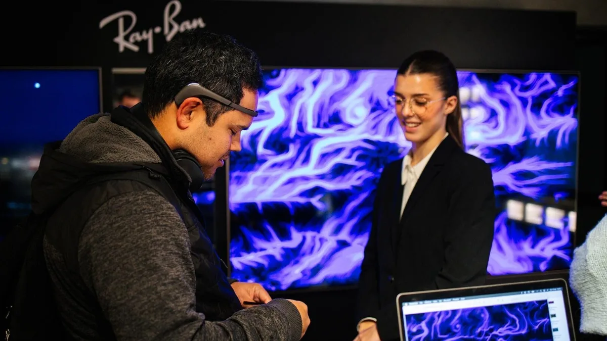 In un ambiente scuro con il logo "Ray-Ban" su una parete, un uomo di profilo con un sensore EEG sulla fronte e cuffie al collo guarda in basso verso le proprie mani. Di fronte a lui una donna in abito scuro e occhiali sorride. Alle loro spalle un grande schermo mostra un'opera dalle linee e dai vortici viola e blu su fondo scuro; in primo piano un portatile aperto che riporta la stessa opera.