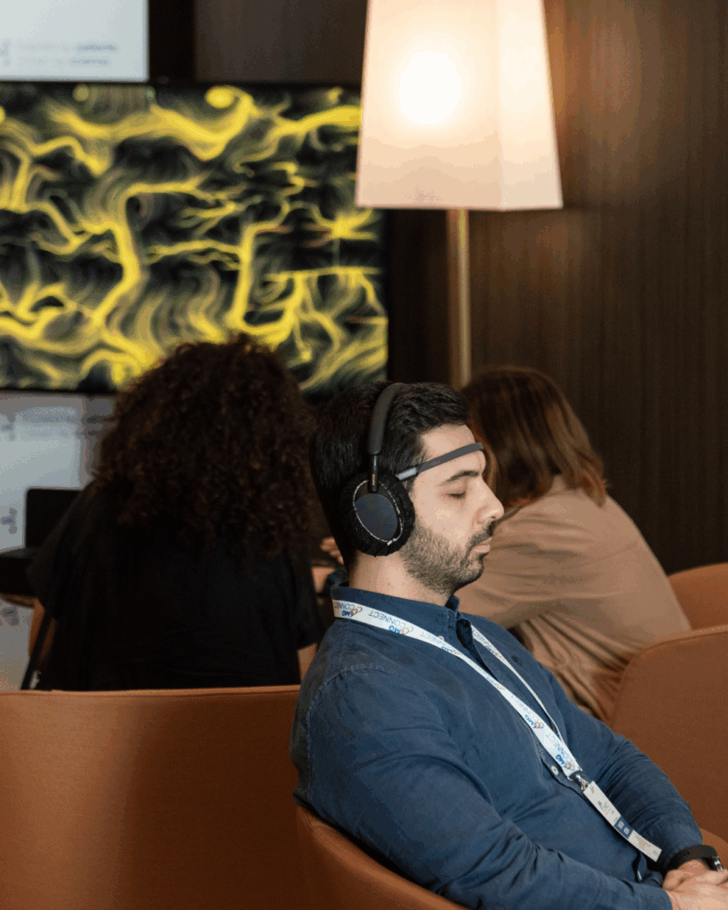 Un uomo seduto in poltrona in un'area lounge, con un sensore EEG sulla fronte e cuffie nere over-ear, tiene gli occhi chiusi. Indossa una camicia blu e un pettorale al collo. Alle sue spalle uno schermo mostra un'opera dalle fitte linee e dai vortici gialli su fondo scuro; a destra una lampada da terra accesa. Sullo sfondo altre persone sedute.