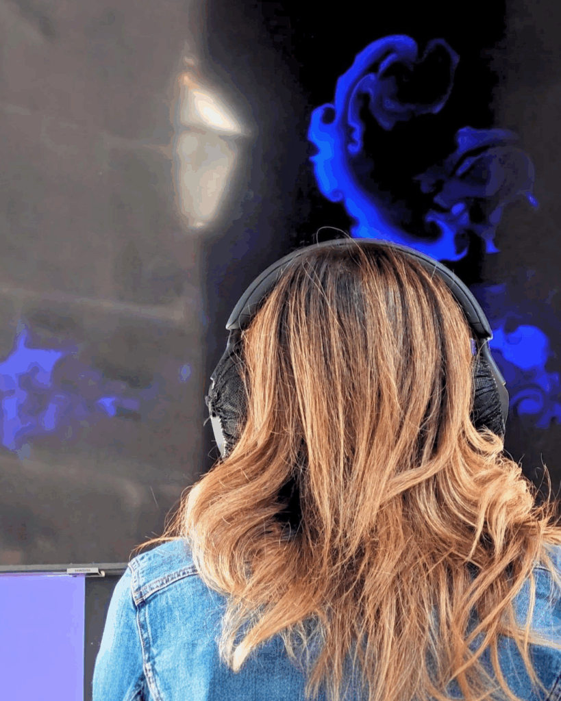 Una donna ripresa di spalle, con capelli biondo ramato, cuffie nere over-ear e giacca di jeans, osserva uno schermo scuro. Sullo schermo compaiono volute e vortici blu su fondo nero, concentrati nella parte alta e con alcune tracce più diffuse ai lati. La superficie dello schermo riflette una luce chiara.