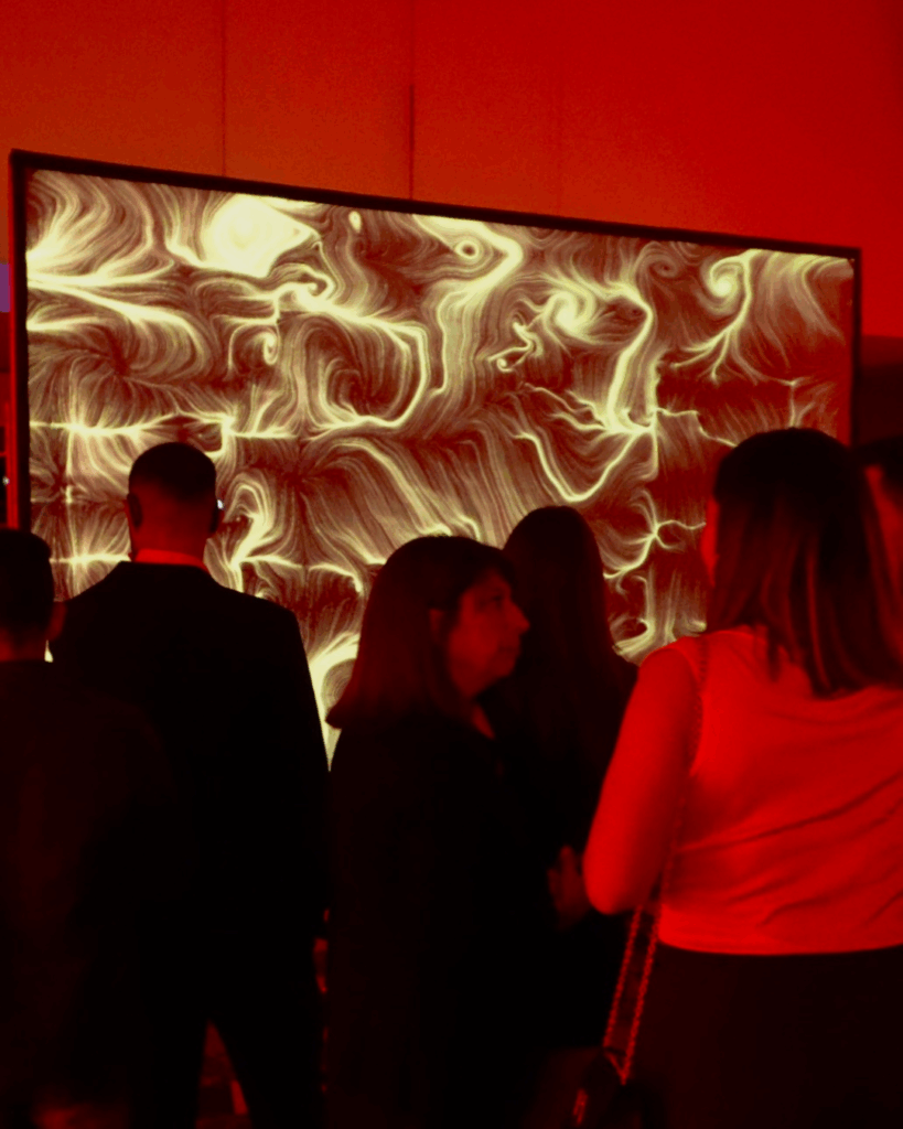 In un ambiente serale illuminato di rosso, un gruppo di persone in piedi, in controluce, osserva e conversa davanti a un grande schermo a parete. Sullo schermo un'opera dalle fitte linee e dai vortici in tonalità chiare e dorate su fondo scuro. Le sagome delle persone si stagliano scure in primo piano.