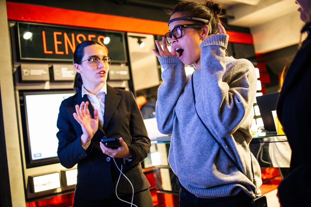 In uno store di occhiali, sotto un'insegna arancione con la scritta "LENSES", una donna con maglione grigio indossa un sensore EEG sulla fronte e occhiali da vista, la bocca aperta e le mani vicine al viso in un gesto di stupore. Accanto, una hostess in abito scuro con uno smartphone in mano le parla, indicandola con l'altra mano. Intorno, espositori di occhiali su pareti scure e rosse.