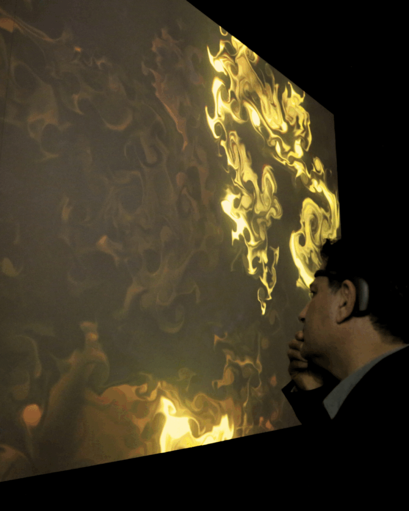 Un uomo di profilo, con un sensore EEG indossato dietro l'orecchio e la mano vicino al mento, osserva una grande proiezione che mostra un'opera dai vortici fluidi dorati e verdi su fondo scuro.