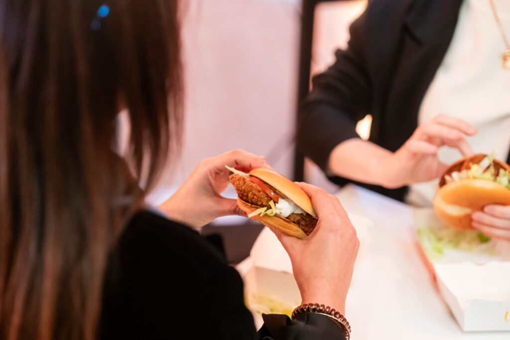 Primo piano di due mani che tengono un panino con una cotoletta impanata, insalata, pomodoro e salsa bianca, sopra una vaschetta di cartone bianca. In primo piano, sfocata di spalle, una persona con capelli lunghi scuri; sulla destra un'altra persona in giacca scura tiene un secondo panino simile su un tavolo chiaro. Lo sfondo è chiaro e sfocato.
