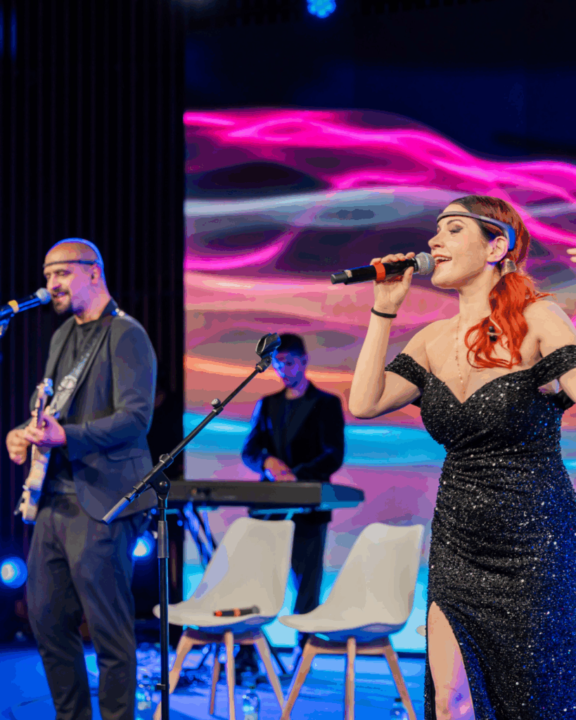 Su un palco, una cantante con abito nero lungo e un sensore EEG sulla fronte canta al microfono. A sinistra un chitarrista, anch'egli con un sensore EEG sul capo, suona una chitarra elettrica; sullo sfondo un tastierista dietro le sedie bianche vuote. Alle loro spalle un grande schermo proietta un'immagine astratta dalle fasce fluide rosa, viola e azzurre. Luci di scena blu illuminano il palco.