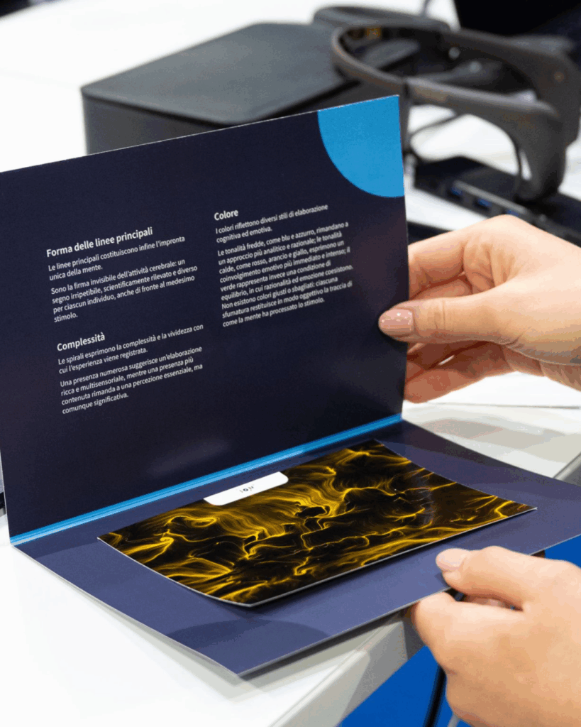 Due mani tengono aperto un pieghevole blu scuro: sulla pagina di sinistra alcuni paragrafi di testo con i titoli "Forma delle linee principali", "Complessità" e "Colore", su quella di destra è applicata la stampa di un'opera dalle fitte linee gialle su fondo nero. Sullo sfondo un piano con un sensore appoggiato.