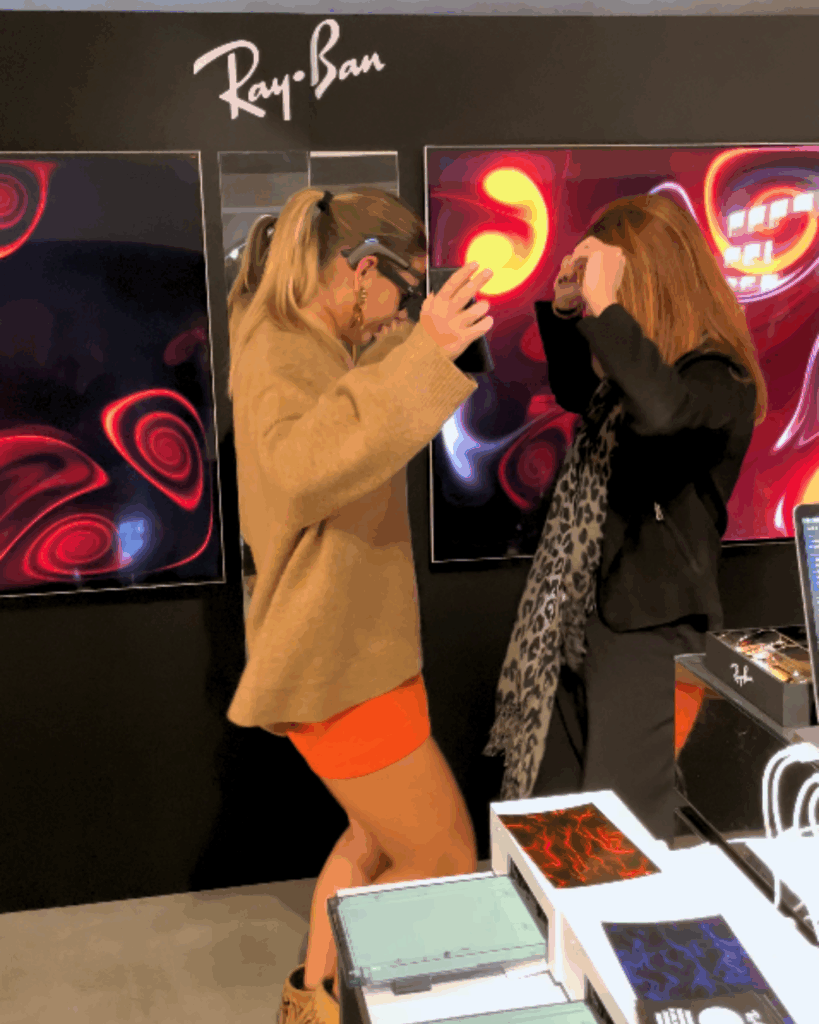 Sotto il logo "Ray-Ban" su una parete scura, due donne interagiscono con gesti animati davanti a due schermi: quello a sinistra mostra un'opera dalle linee e dai vortici blu e rossi su fondo scuro, quello a destra un'opera dai vortici rossi e gialli. In primo piano un espositore con stampe e materiali dalle grafiche colorate. Una delle due indossa un sensore EEG sulla fronte.