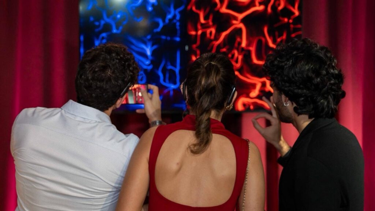 Tre persone di spalle osservano due opere BrainArt®, una blu e una rossa, davanti a tende di velluto rosso. Uno di loro fotografa con lo smartphone.