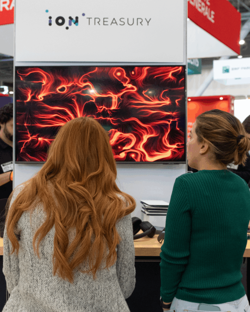 Due donne di spalle osservano lo schermo dello stand ION Treasury, che mostra un'opera dalle linee e dai vortici rossi e arancioni su fondo nero; intorno, i padiglioni e le insegne di altri stand fieristici.