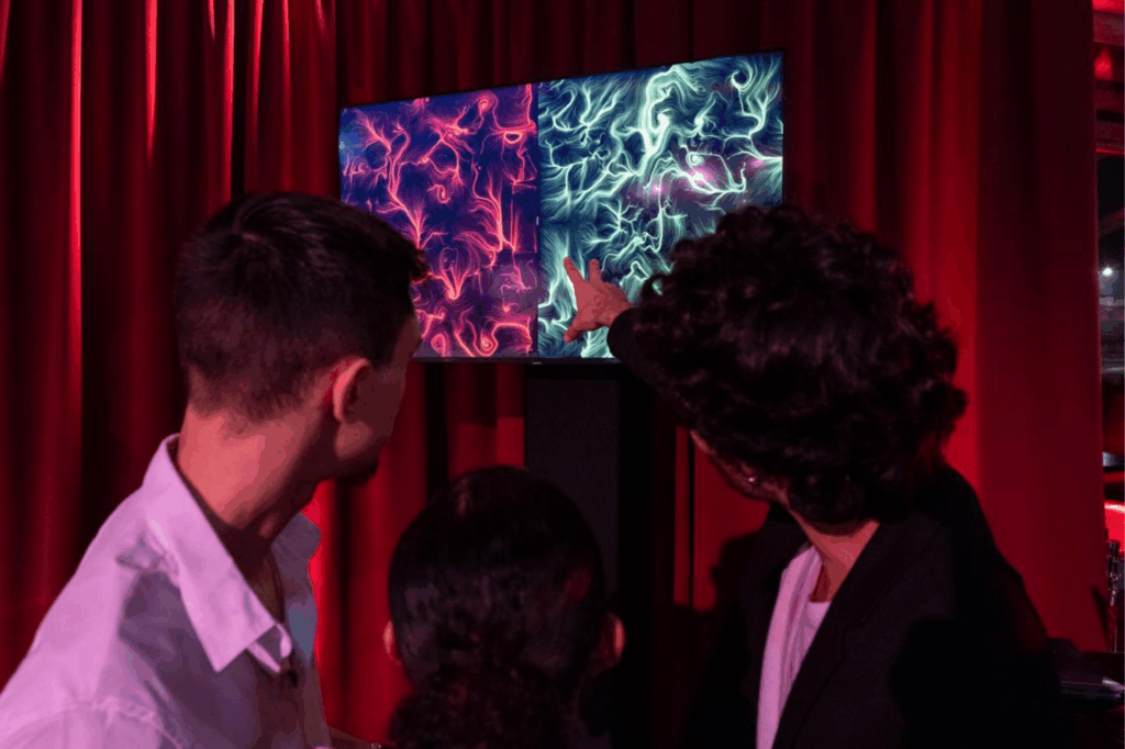 Tre persone osservano uno schermo con due opere BrainArt®, una rossa e una verde generate dalle onde cerebrali, appese davanti a una tenda di velluto rosso.