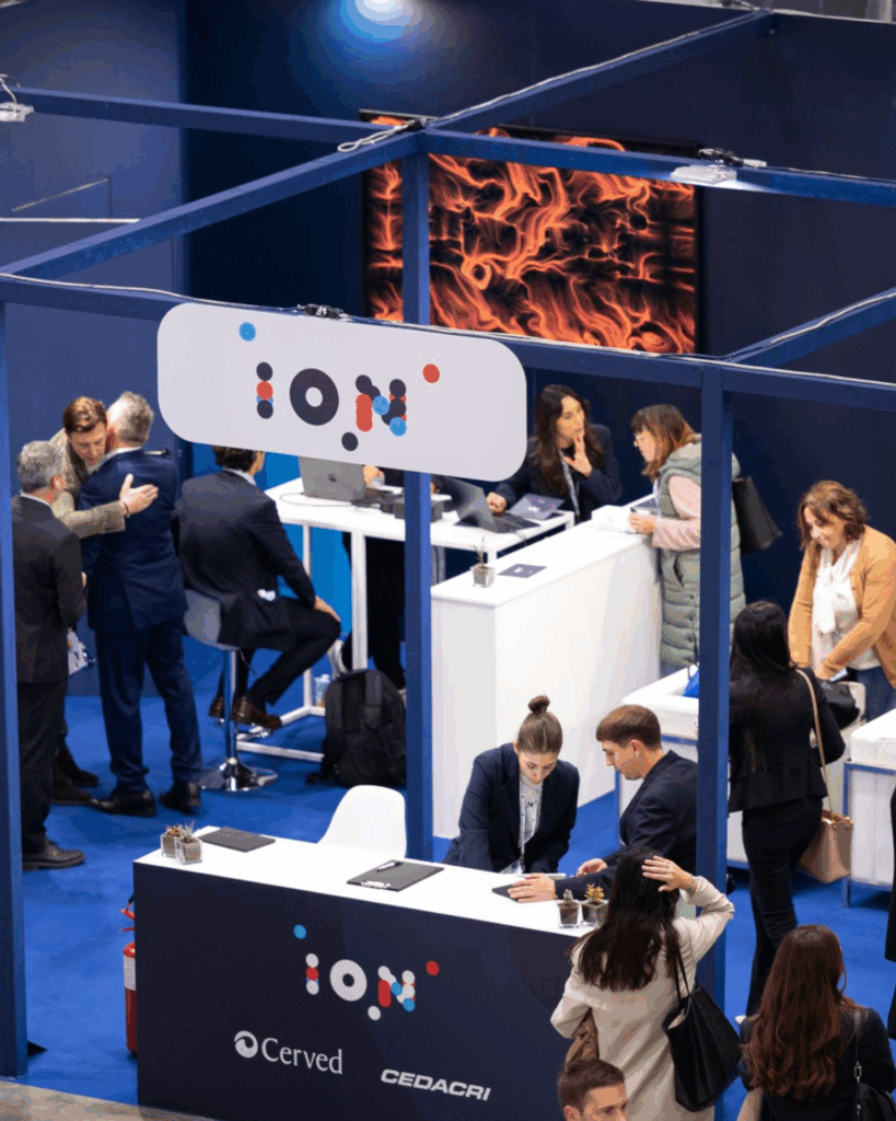Vista dall'alto di uno stand fieristico blu con il logo ION: sulla parete di fondo uno schermo mostra un'opera dalle linee e dai vortici arancioni; diverse persone conversano ai banconi, uno dei quali riporta i loghi Cerved e CEDACRI.