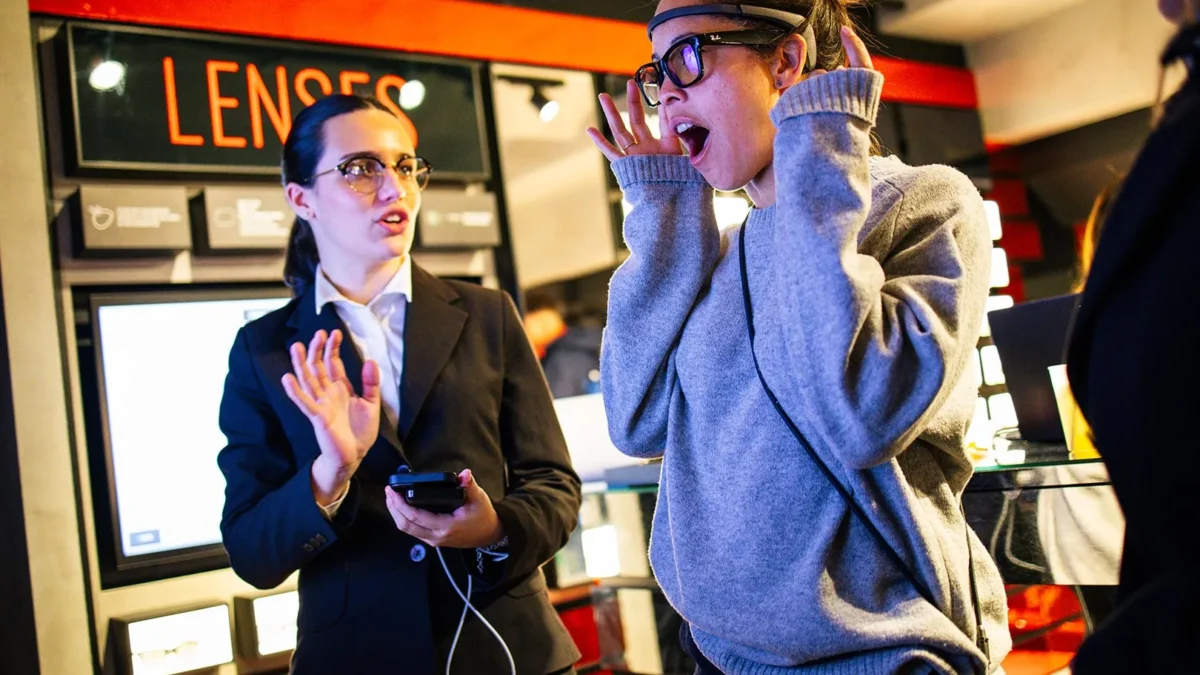 In uno store di occhiali, sotto un'insegna arancione con la scritta "LENSES", una donna con maglione grigio indossa un sensore EEG sulla fronte e occhiali da vista, la bocca aperta e le mani vicine al viso in un gesto di stupore. Accanto, una hostess in abito scuro con uno smartphone e un auricolare con cavo le parla, indicandola con l'altra mano. Intorno, espositori di occhiali illuminati su pareti scure e rosse.