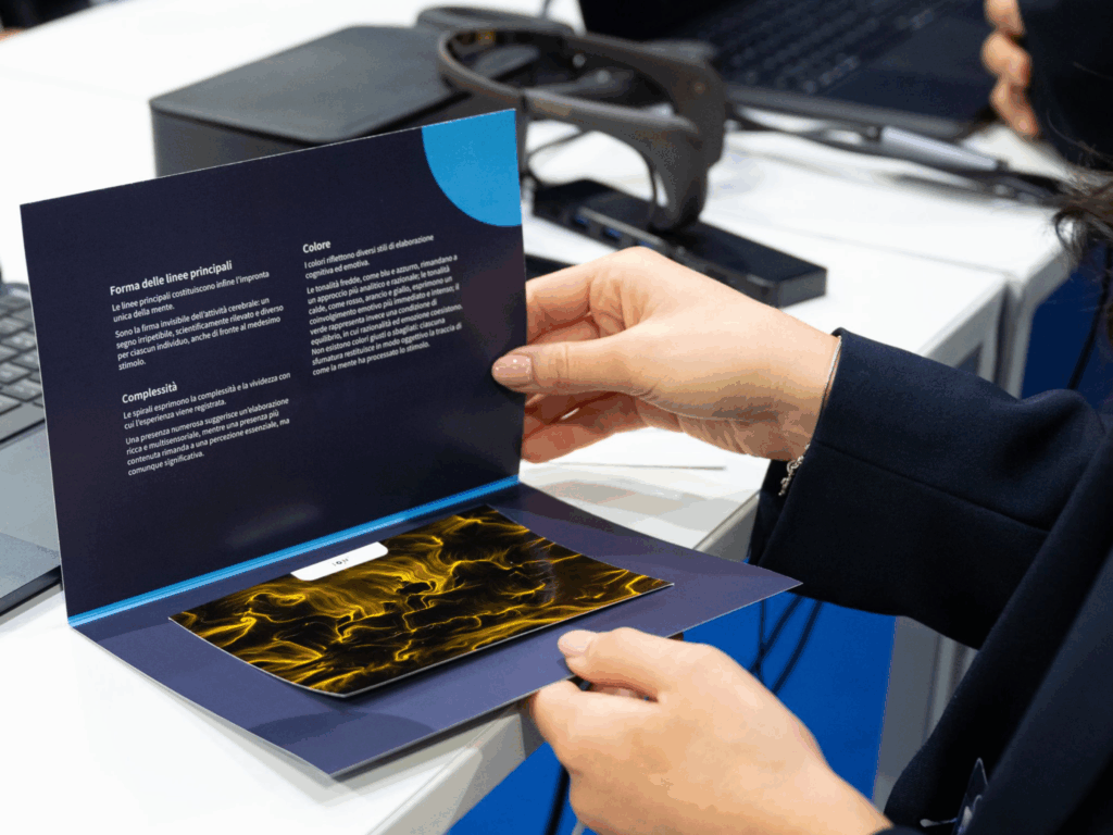 Due mani aprono un pieghevole blu scuro: nell'anta superiore tre paragrafi di testo intitolati "Forma delle linee principali", "Complessità" e "Colore"; nell'anta inferiore la stampa di un'opera dalle linee e dai vortici gialli su fondo nero. Sullo sfondo, appoggiato al bancone, un sensore EEG.