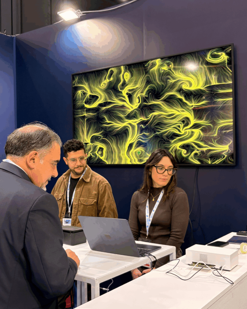 Un uomo in giacca, di profilo, guarda lo schermo di un computer portatile appoggiato sul bancone di uno stand; dietro di lui due persone con pass al collo seguono la scena, davanti a uno schermo che mostra un'opera dalle linee e dai vortici gialli.