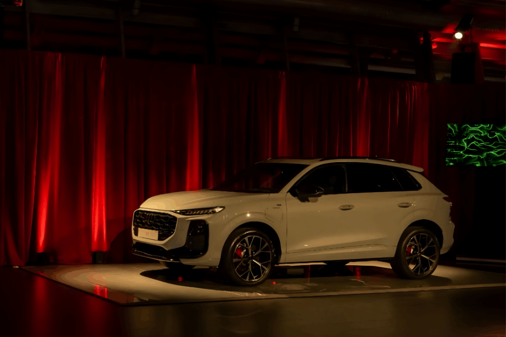 SUV Audi Q3 bianco esposto su una pedana illuminata davanti a tende di velluto rosso, con un quadro BrainArt verde su uno schermo a destra.