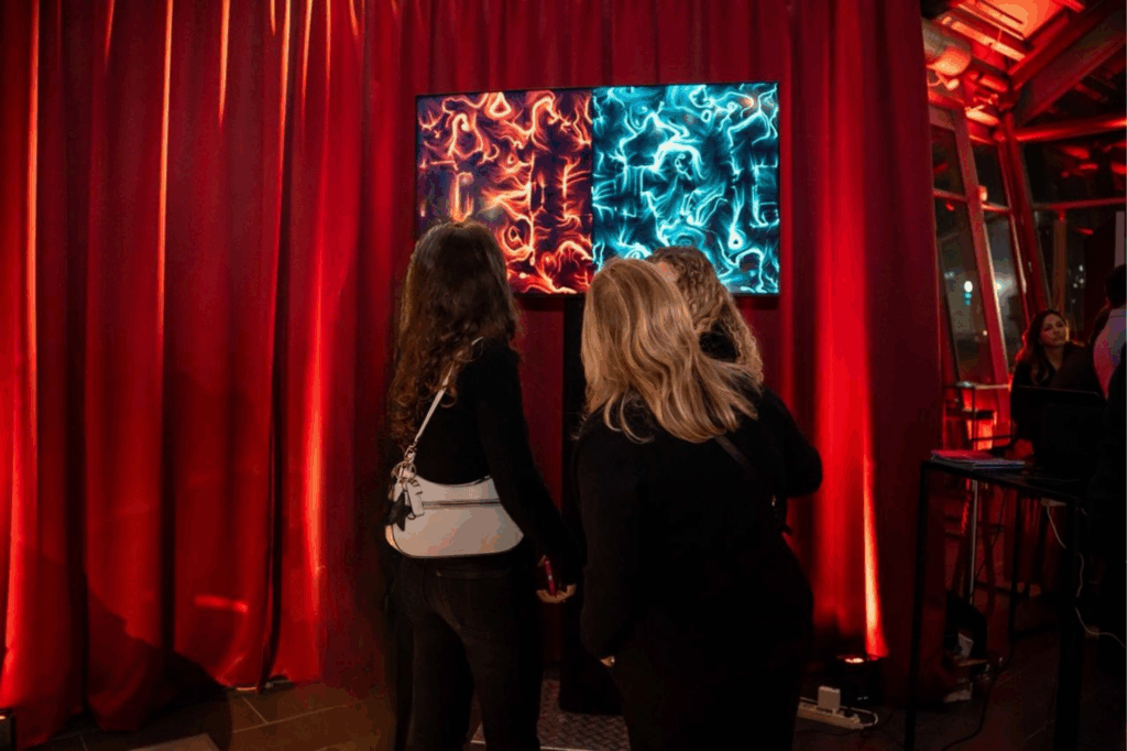 Tre donne di spalle osservano uno schermo con due opere BrainArt®, una arancione e una blu, posizionato davanti a una tenda di velluto rosso