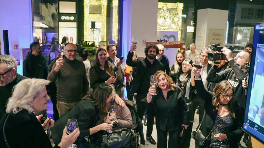 Foto di gruppo di partecipanti a un evento serale, molti con un calice in mano, davanti a uno schermo con un quadro BrainArt® rosso e un pannello con il logo AISLA