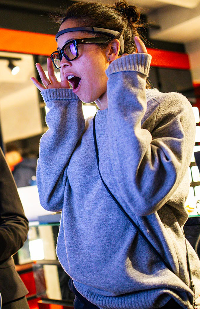 In uno store di occhiali, sotto un'insegna arancione con la scritta "LENSES", una donna con maglione grigio indossa un sensore EEG sulla fronte e occhiali da vista, la bocca aperta e le mani vicine al viso in un gesto di stupore. Accanto, una hostess in abito scuro con uno smartphone in mano le parla, indicandola con l'altra mano. Intorno, espositori di occhiali su pareti scure e rosse.