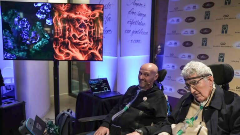 Due uomini in carrozzina a un evento, uno con un dispositivo EEG indossato, davanti a uno schermo con due quadri BrainArt® blu e rosso e un pannello con il logo AISLA