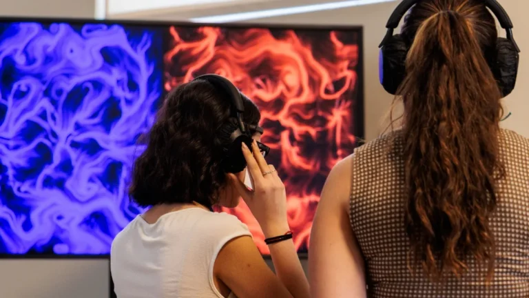 Due ragazze riprese di spalle indossano cuffie nere over-ear e osservano due schermi affiancati: quello a sinistra mostra un'opera dalle volute e dai vortici blu e viola su fondo scuro, quello a destra un'opera dalle volute e dai vortici rossi. La ragazza a sinistra, in canotta bianca, si tiene una mano sulla cuffia; quella a destra indossa un top a quadretti. L'ambiente è chiaro e illuminato.