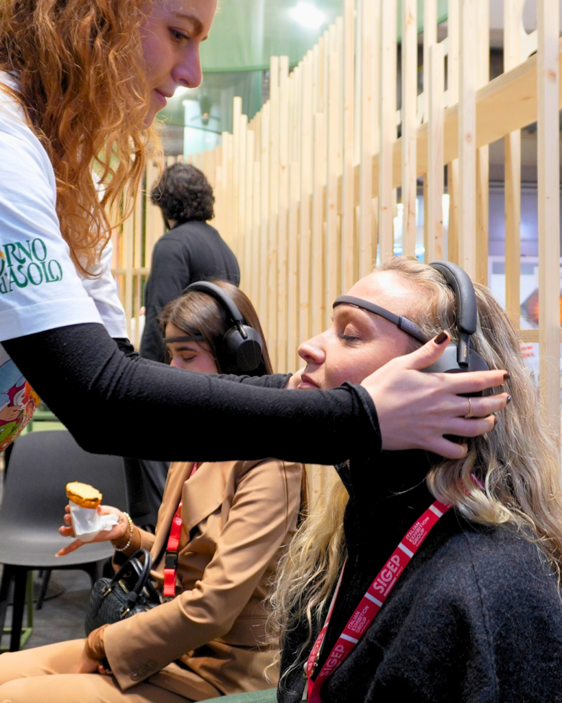 Una ragazza con maglietta Forno d'Asolo sistema con una mano le cuffie e il sensore EEG sulla testa di una visitatrice seduta a occhi chiusi con il pass SIGEP al collo, mentre nell'altra mano tiene un cornetto in un tovagliolo; accanto, un'altra visitatrice seduta con le cuffie.