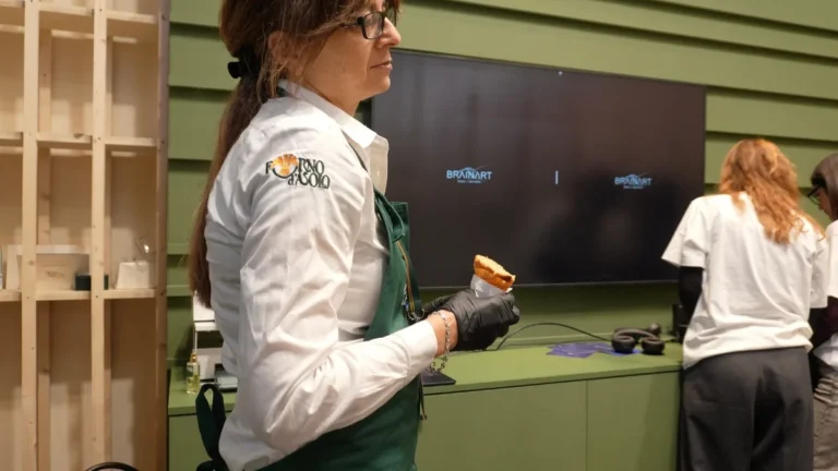 Una donna con occhiali, camicia bianca con il logo Forno d'Asolo e grembiule verde tiene in mano, con un guanto nero, un cornetto avvolto in un tovagliolo; alle sue spalle una parete verde a doghe con uno schermo che mostra due loghi BrainArt® affiancati su fondo scuro, un piano con delle cuffie appoggiate e, sulla destra, due persone in maglietta bianca di spalle.