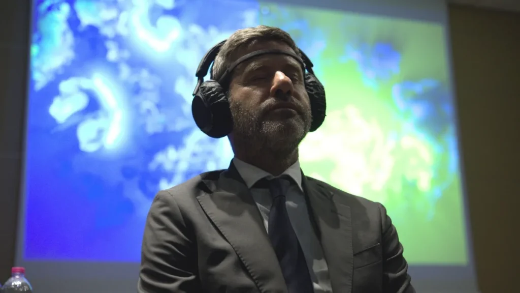 Un uomo in giacca e cravatta scure, con il sensore appoggiato sulla fronte e le cuffie sulle orecchie, ascolta a occhi chiusi; alle sue spalle una grande proiezione mostra un'opera dalle fitte linee e dai vortici azzurri e verdi su fondo scuro.