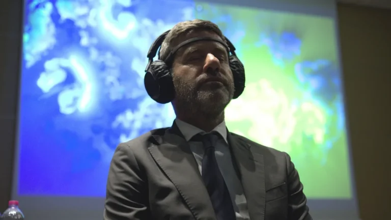 Un uomo in giacca e cravatta scure, con il sensore appoggiato sulla fronte e le cuffie sulle orecchie, ascolta a occhi chiusi; alle sue spalle una grande proiezione mostra un'opera dalle fitte linee e dai vortici azzurri e verdi su fondo scuro.