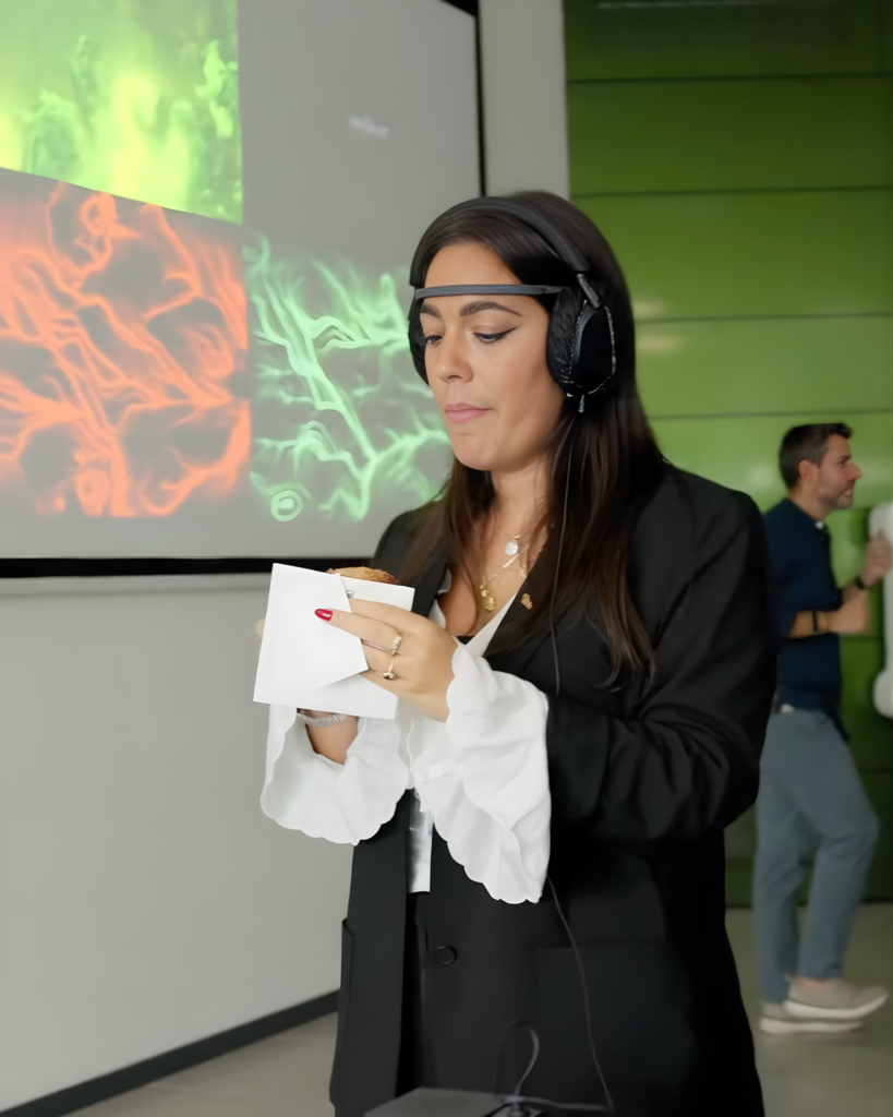 Una donna con un sensore EEG sulla fronte e le cuffie in testa tiene tra le mani un cornetto avvolto in un tovagliolo bianco e lo guarda; alle sue spalle una proiezione con opere dalle linee e dai vortici arancioni e verdi.
