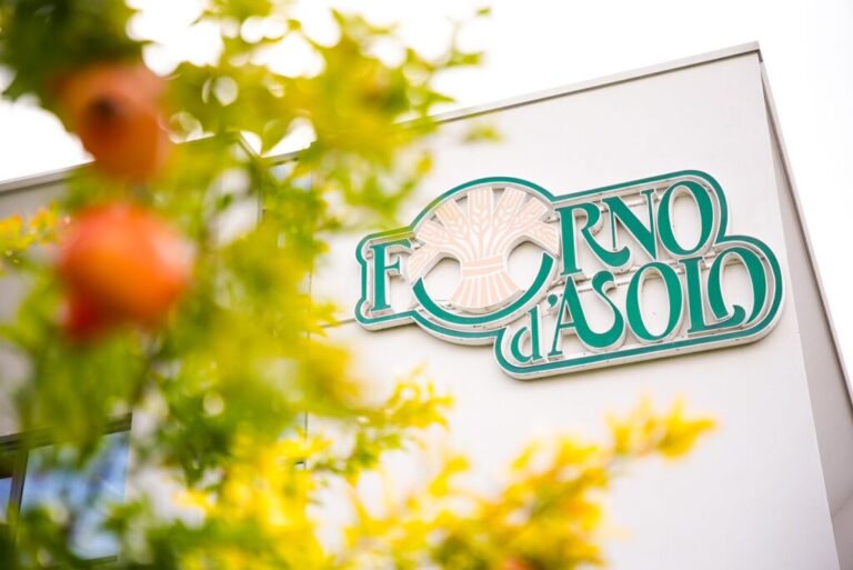 Insegna con il logo Forno d'Asolo, in verde e rosa con un covone di grano, montata sulla facciata bianca di un edificio; in primo piano, sfocati, rami con foglie verdi e gialle e frutti rossi.