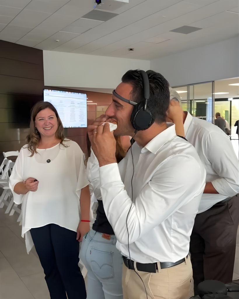 Un uomo con un sensore EEG sulla fronte e le cuffie in testa assaggia un cornetto tenuto in un tovagliolo; accanto a lui una donna sorride, mentre sullo sfondo altre persone vivono la stessa esperienza.