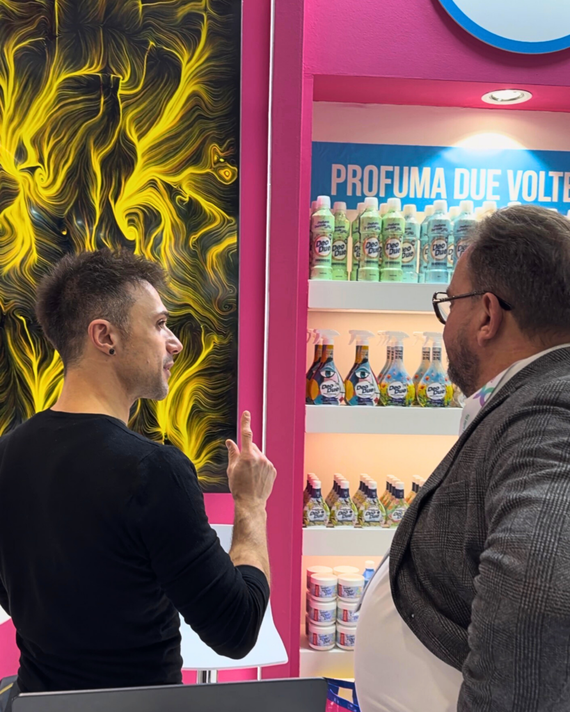 A uno stand fieristico, due uomini di profilo dialogano; quello a sinistra, in maglia scura, indica con un dito verso l'alto. Alle loro spalle, a sinistra, uno schermo verticale mostra un'opera dalle fitte linee e dai vortici gialli su fondo scuro. A destra, scaffali rosa con flaconi e spray di prodotti per la casa e il claim "Profuma due volt…". Uno dei due porta un pettorale al collo.