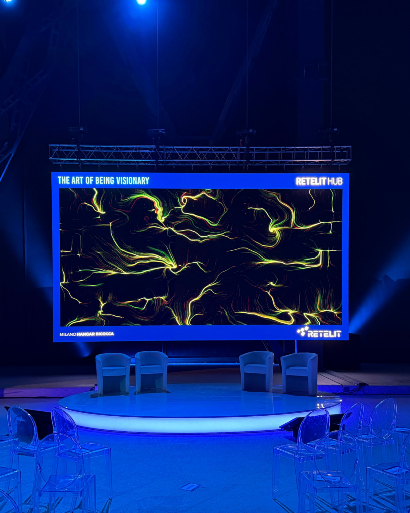 Palco vuoto con poltroncine e un maxischermo che mostra un'opera dalle linee e dai vortici gialli e verdi su fondo nero, incorniciata dal claim "The Art of Being Visionary" e dai loghi Retelit Hub e Retelit; la sala è illuminata di blu.