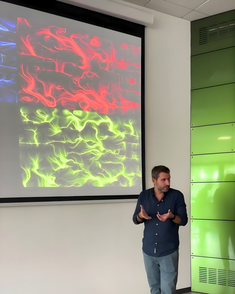Un uomo in piedi parla gesticolando accanto a uno schermo di proiezione che mostra due opere affiancate, una dalle forme fluide rosse e una dalle linee e dai vortici verdi; sulla destra una parete verde.