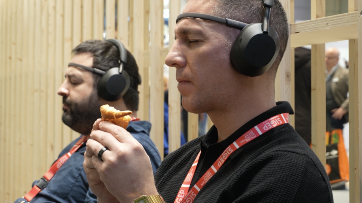 Due uomini seduti uno accanto all'altro, ciascuno con un sensore EEG sulla fronte e le cuffie in testa e con il pass SIGEP al collo: quello in primo piano tiene in mano un cornetto già addentato e tiene gli occhi socchiusi; alle loro spalle, la parete in listelli di legno dello stand.