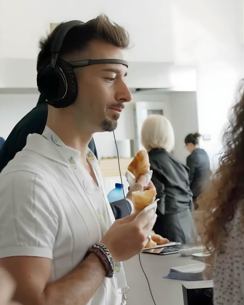 Un uomo di profilo, con un sensore EEG sulla fronte e le cuffie in testa, tiene in mano un cornetto avvolto in un tovagliolo; sullo sfondo altre persone in piedi accanto a un banco.