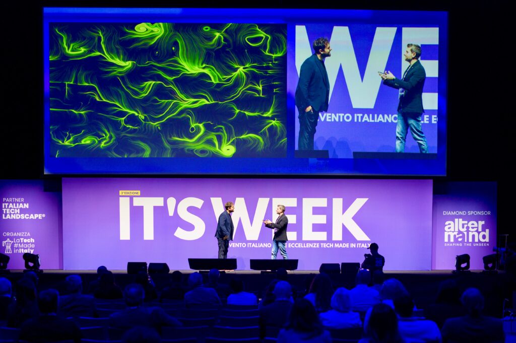 Due uomini in piedi sul palco di un evento tech si parlano davanti a un fondale con la scritta "IT'S WEEK"; sopra, un maxischermo diviso mostra a sinistra un'opera dalle linee e dai vortici verdi e a destra la ripresa dal vivo di uno dei due.