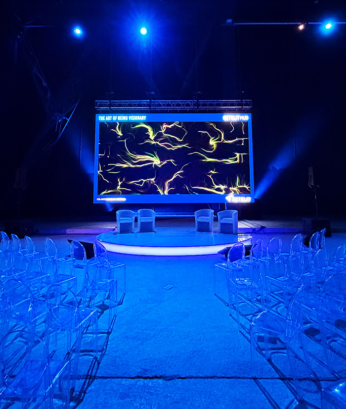 Palco allestito per una live demo BrainArt® con maxischermo che mostra un'opera visiva generata da onde cerebrali davanti a una platea